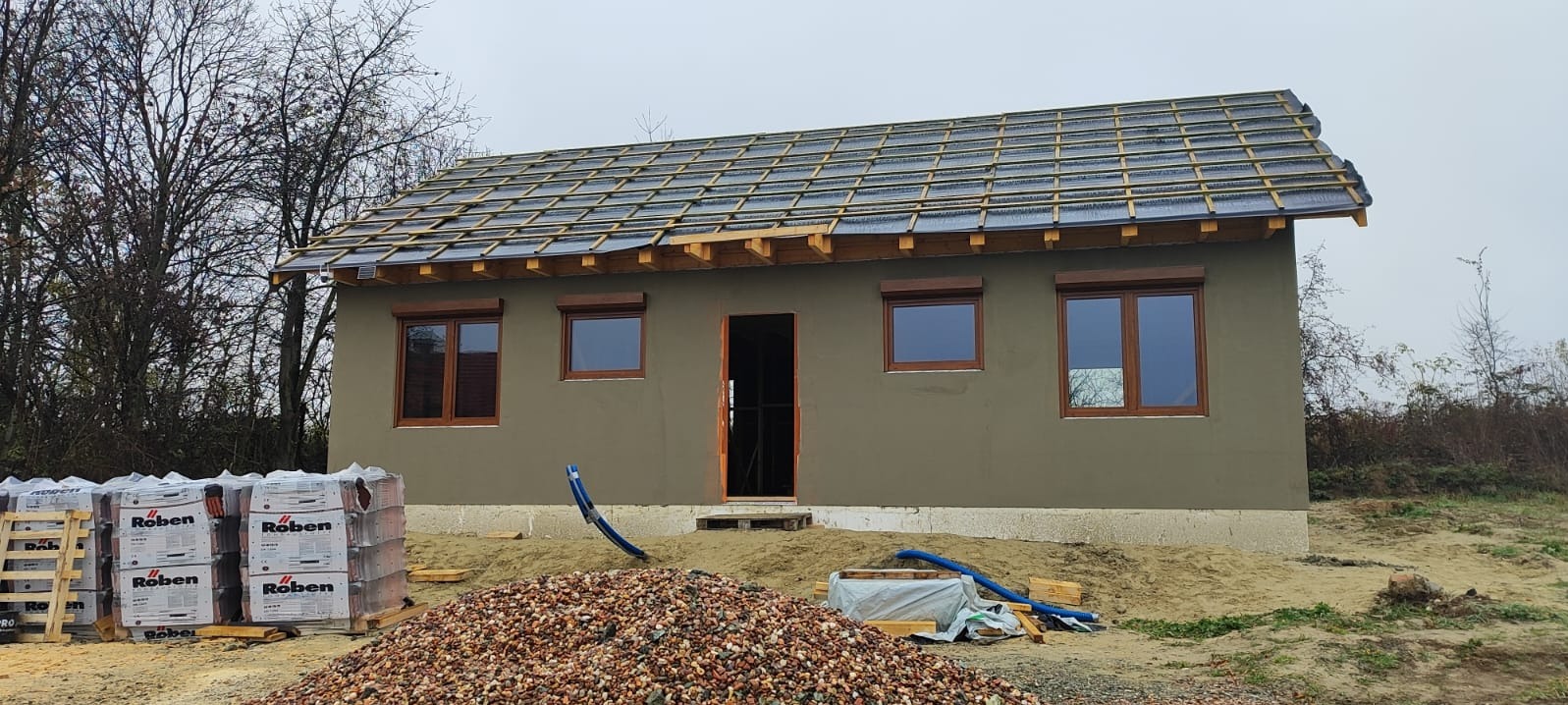 Niedokończony dom z szarą elewacją, drewnianymi oknami i przygotowaną więźbą dachową. Na pierwszym planie sterta kamieni i palety z dachówkami Roben.
