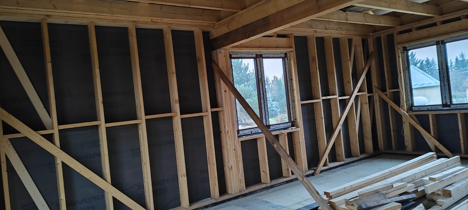 Wnętrze domu w trakcie budowy szkieletowej. Widoczne drewniane ramy ścian z izolacją, okna z widokiem na zewnątrz i stos drewna na podłodze.