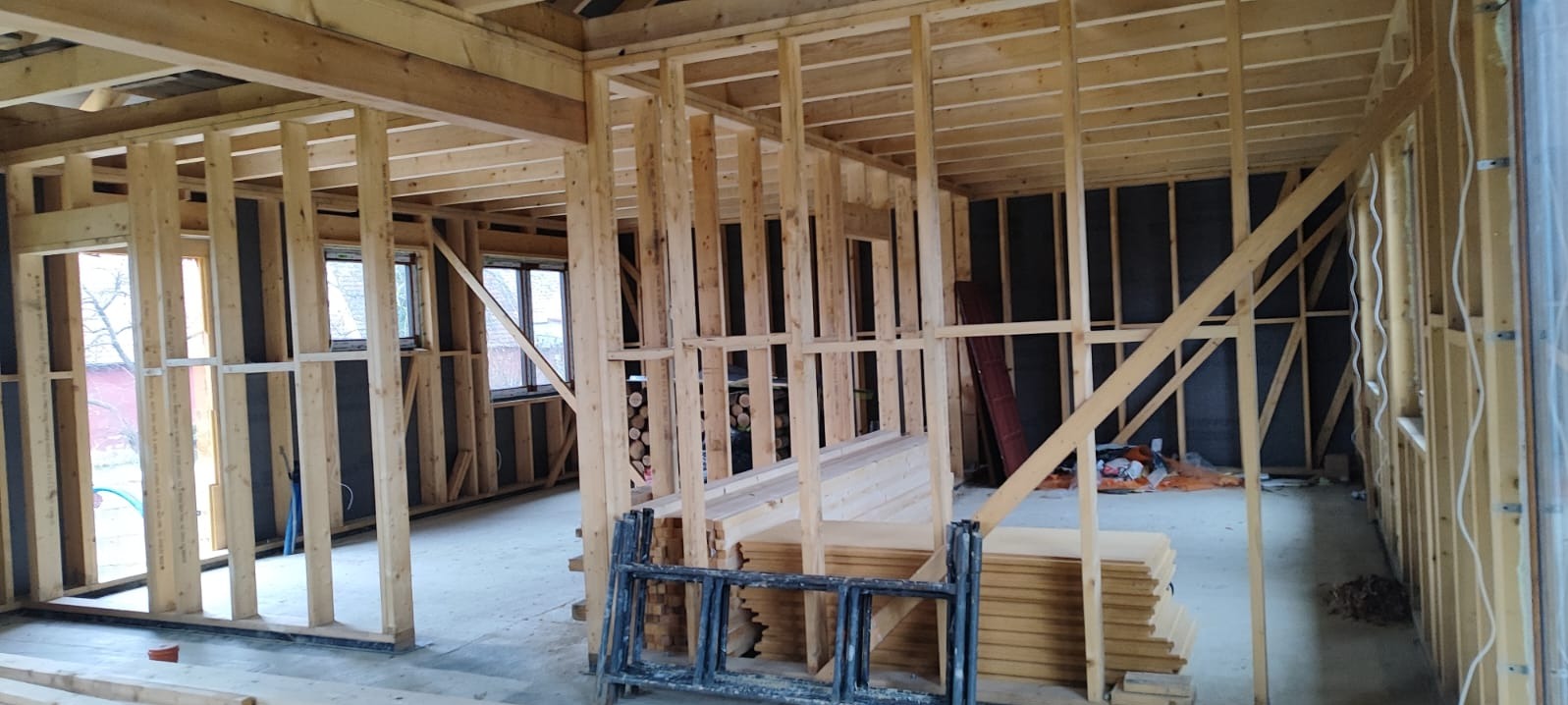 Wnętrze domu w trakcie budowy w technologii szkieletowej. Widoczne drewniane ramy ścian, okna, izolacja, stos desek i drabina. Surowy stan, etap wznoszenia konstrukcji.