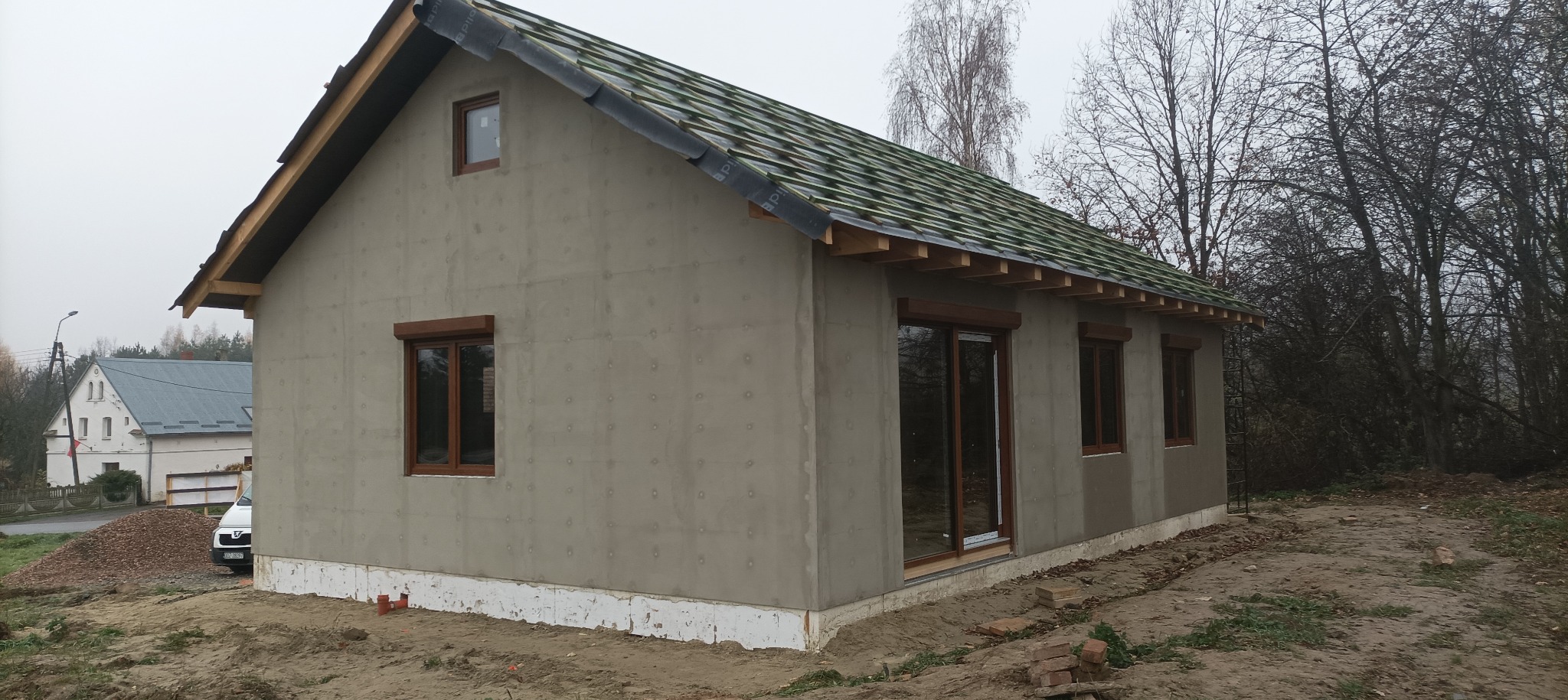 Dom szkieletowy w trakcie budowy: ocieplone ściany, drewniana konstrukcja dachu z zieloną membraną. Widoczne okna z brązowymi ramami i fragment otoczenia.