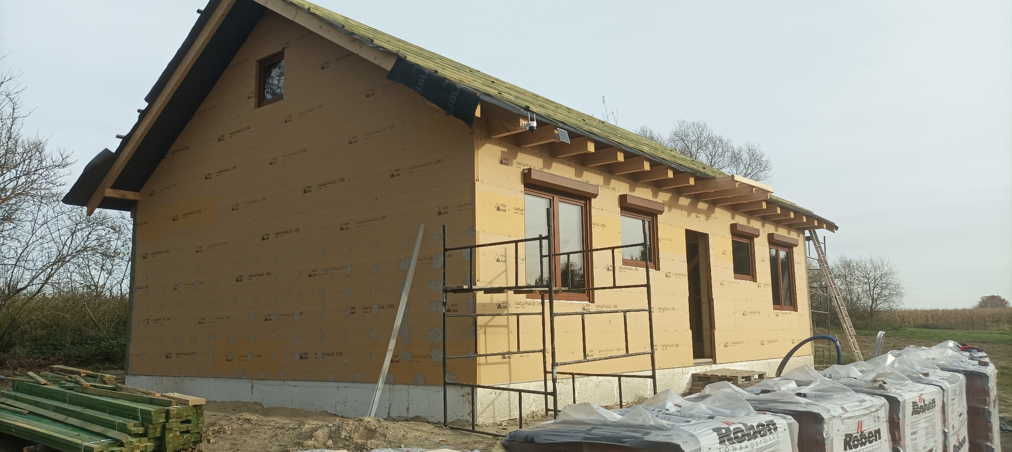 Budowa domu szkieletowego w trakcie: widoczne ściany z płyt, okna z roletami zewnętrznymi, rusztowanie i materiały budowlane. Dach w trakcie wykończenia.