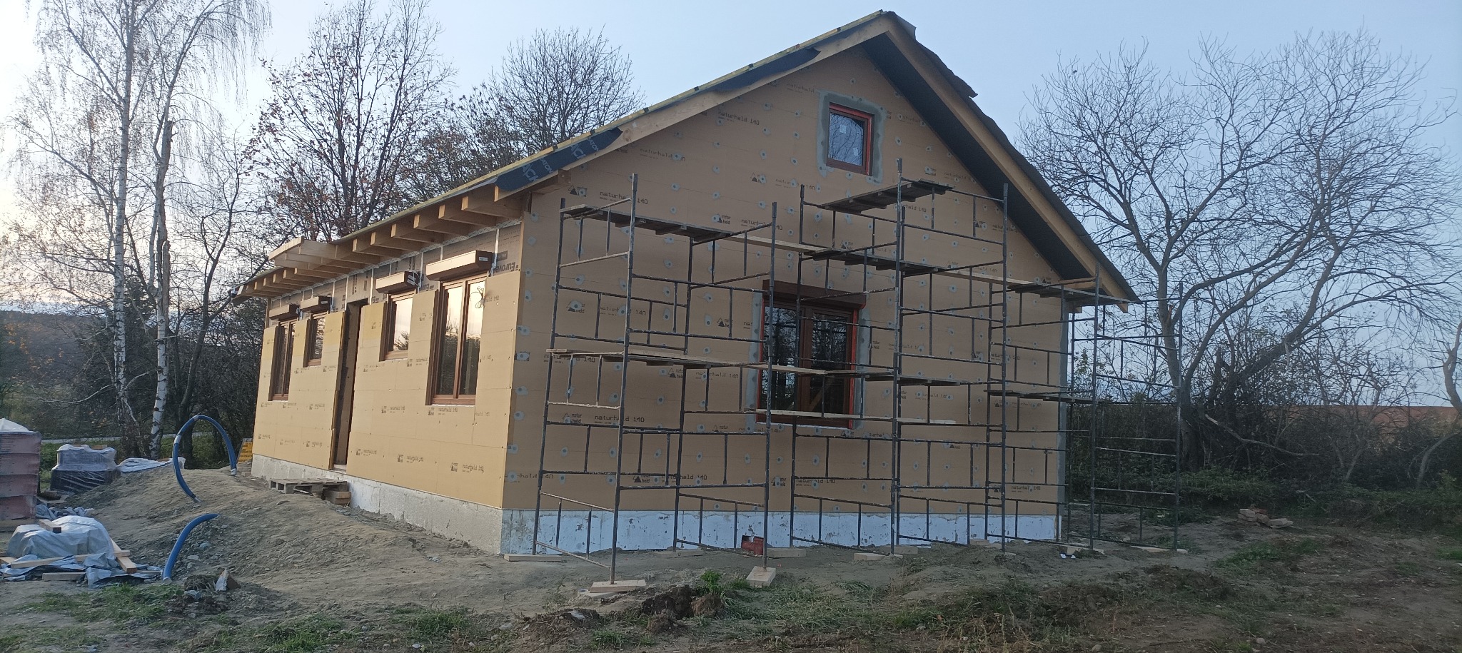 Surowy dom szkieletowy w trakcie budowy, z widocznymi rusztowaniami i oknami z brązowymi ramami, na tle drzew bez liści i pochmurnego nieba.