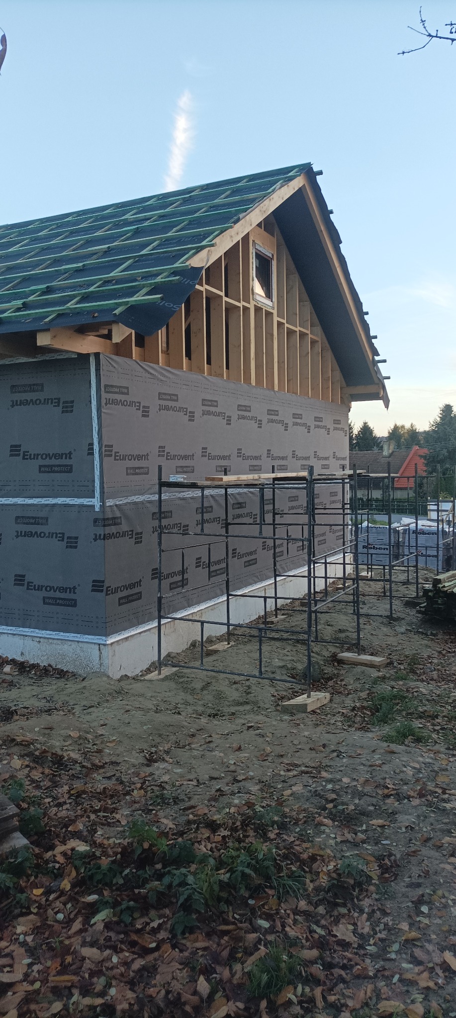 Budowa domu szkieletowego w trakcie: widoczna konstrukcja, izolacja Eurovent, rusztowania. Ujęcie z dołu, w tle inne budynki i drzewa. Jesienne liście na ziemi.