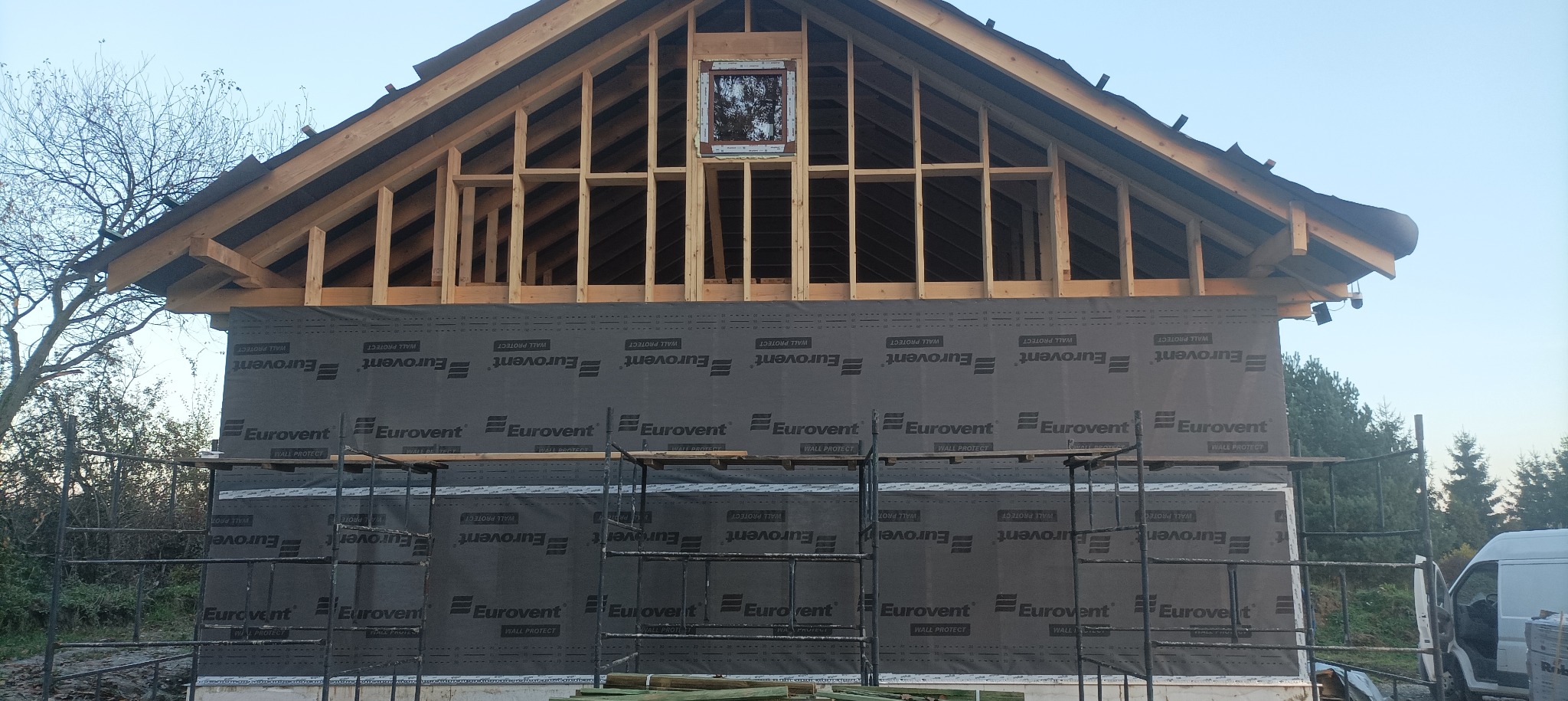 Budowa domu szkieletowego: widok na drewnianą konstrukcję dachu i ściany z membraną Eurovent, rusztowania w tle, etap prac przed wykończeniem elewacji.