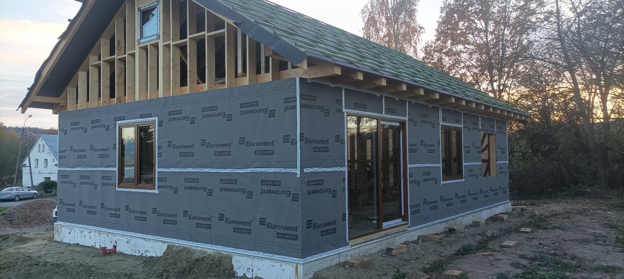 Budowa domu szkieletowego w trakcie: widoczne drewniane elementy konstrukcyjne, ściany pokryte membraną Eurovent, okna wstawione. Etap prac przed ociepleniem i wykończeniem.