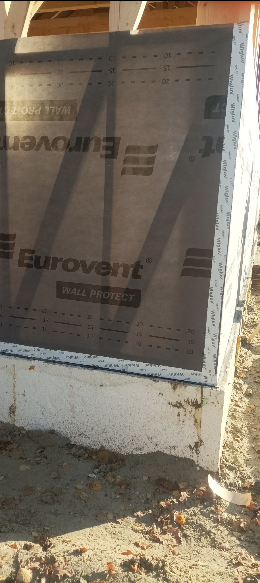 Fragment ściany domu szkieletowego w trakcie budowy, z widoczną membraną Eurovent Wall Protect i taśmą uszczelniającą Wigluv, na fundamencie. Detal izolacji.
