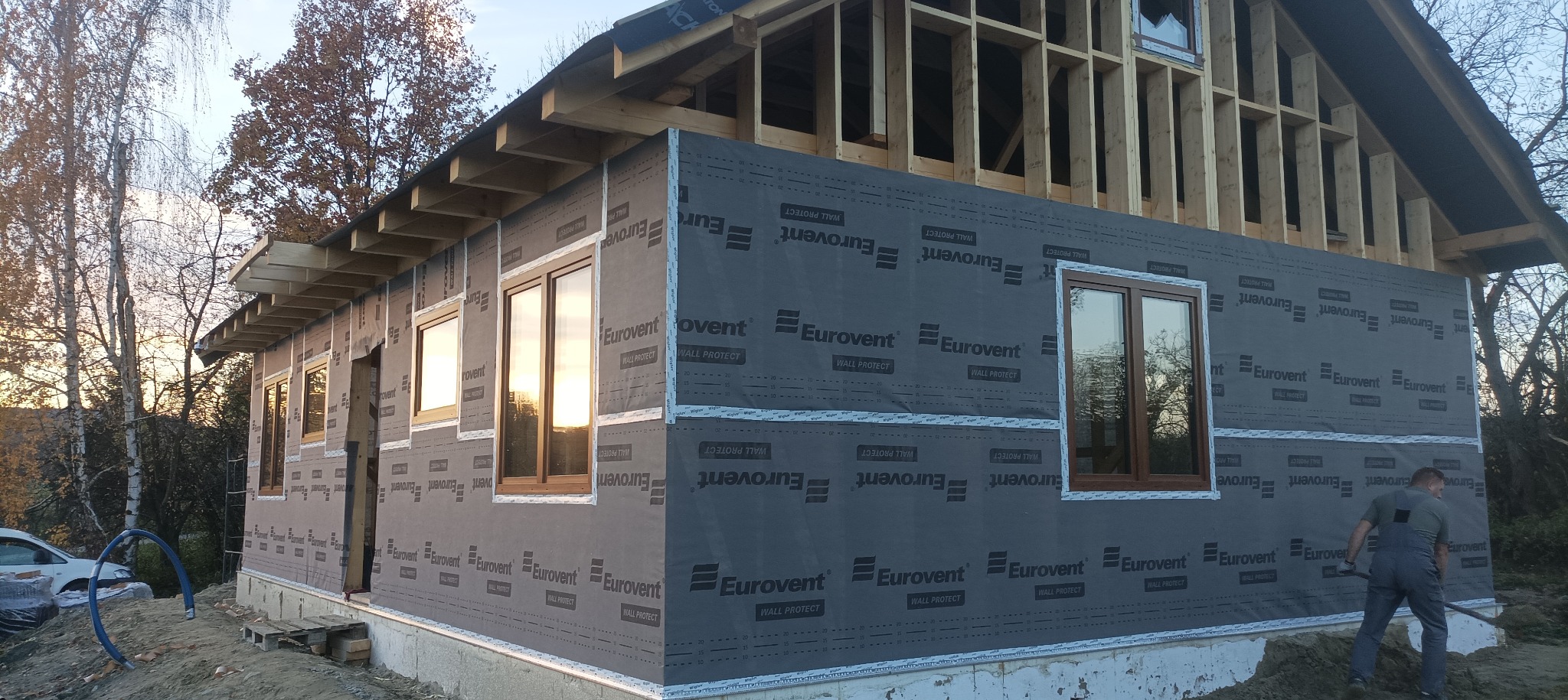 Budowa domu szkieletowego: widok na ścianę z oknami, pokrytą membraną Eurovent, z widoczną konstrukcją więźby dachowej i pracującym mężczyzną w tle.