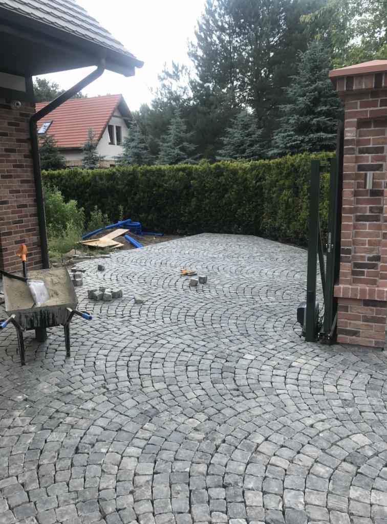 Fragment podjazdu z szarej kostki granitowej ułożonej w okrągły wzór; w tle widoczny wózek budowlany z zaprawą, brama wjazdowa i zadbany żywopłot.
