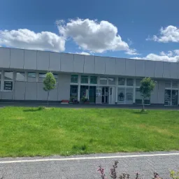 Budynek z napisem 'APTEKA' nad wejściem, przed nim trawnik i dwa małe drzewka. Na oknach widoczne plakaty, w tym jeden z ofertą treningów personalnych i grupowych.