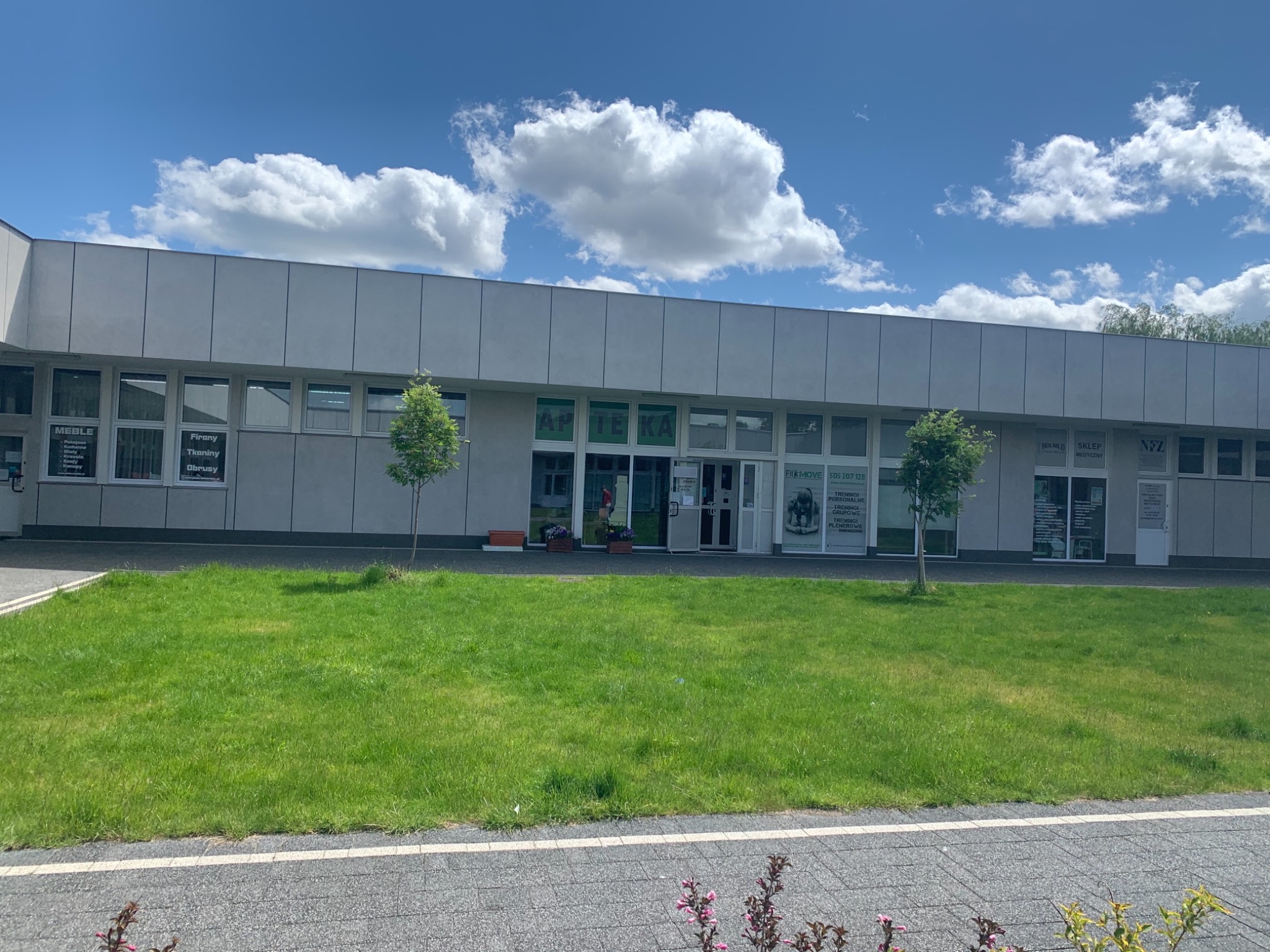Budynek z napisem 'APTEKA' nad wejściem, przed nim trawnik i dwa małe drzewka. Na oknach widoczne plakaty, w tym jeden z ofertą treningów personalnych i grupowych.