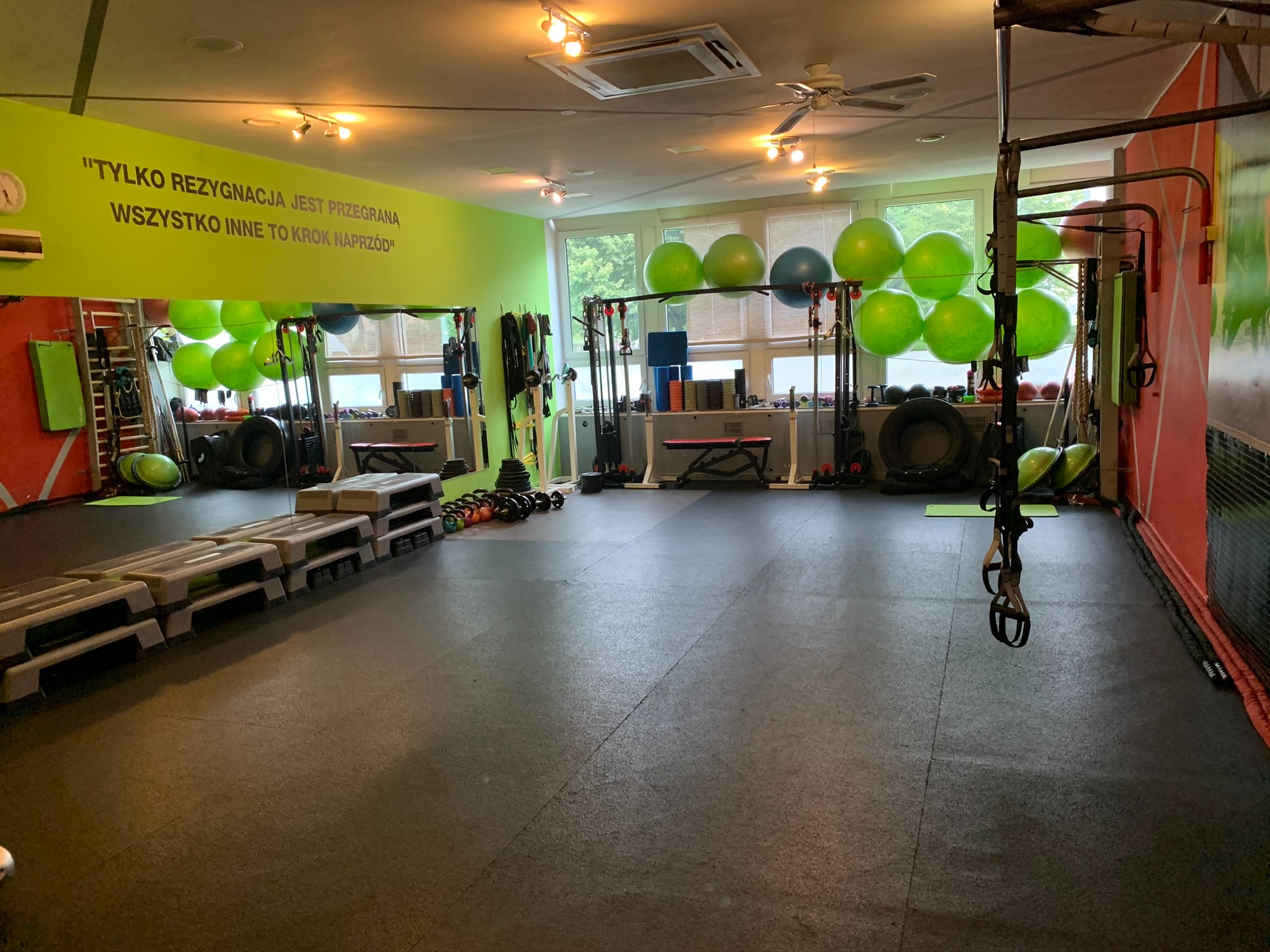 Przestronna, dobrze wyposażona siłownia z zieloną ścianą z motywacyjnym cytatem, sprzętem do ćwiczeń siłowych i cardio, oraz rzędem piłek gimnastycznych ustawionych wzdłuż okien.