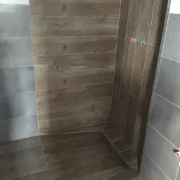 Wykończona kabina prysznicowa z brązowymi płytkami imitującymi drewno na ścianach i podłodze, kontrastującymi z szarymi płytkami na sąsiedniej ścianie, widoczny odpływ liniowy w rogu.