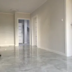 Pok&oacute;j po remoncie z błyszczącą, szarą podłogą z płytek, świeżo pomalowane ściany w odcieniach szarości, białe drzwi wewnętrzne z poziomymi paskami i widocznymi oprawami oświetleniowymi na suficie.