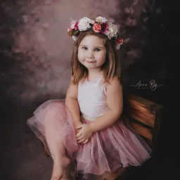 Dziewczynka w różowej tiulowej spódniczce i wianku z kwiatów na głowie, siedząca na drewnianym krześle na tle abstrakcyjnego brązowego tła.