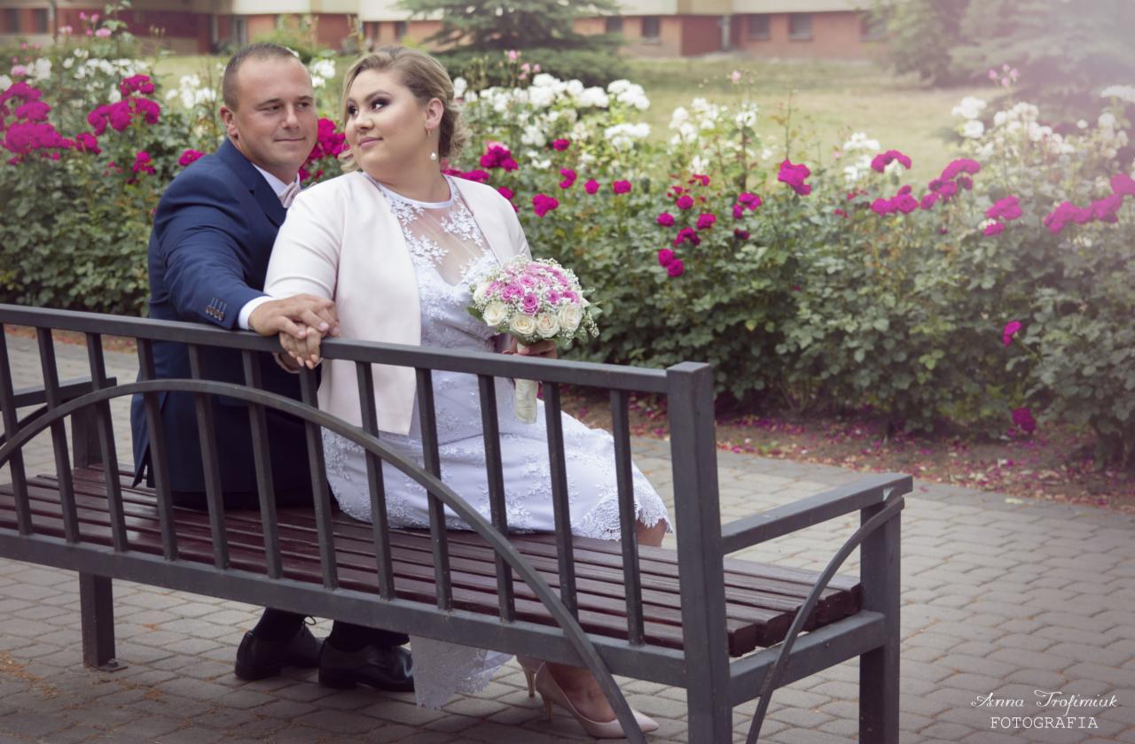Para młoda siedząca na ławce w parku, trzymająca się za ręce, z bukietem kwiatów na tle kwitnących krzewów róż.