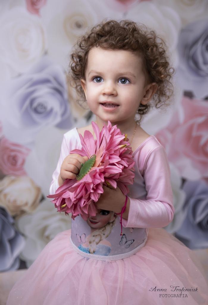 Portret uśmiechniętej dziewczynki z kręconymi włosami, trzymającej duży, różowy kwiat na tle kwiatowej ściany w studio fotograficznym.