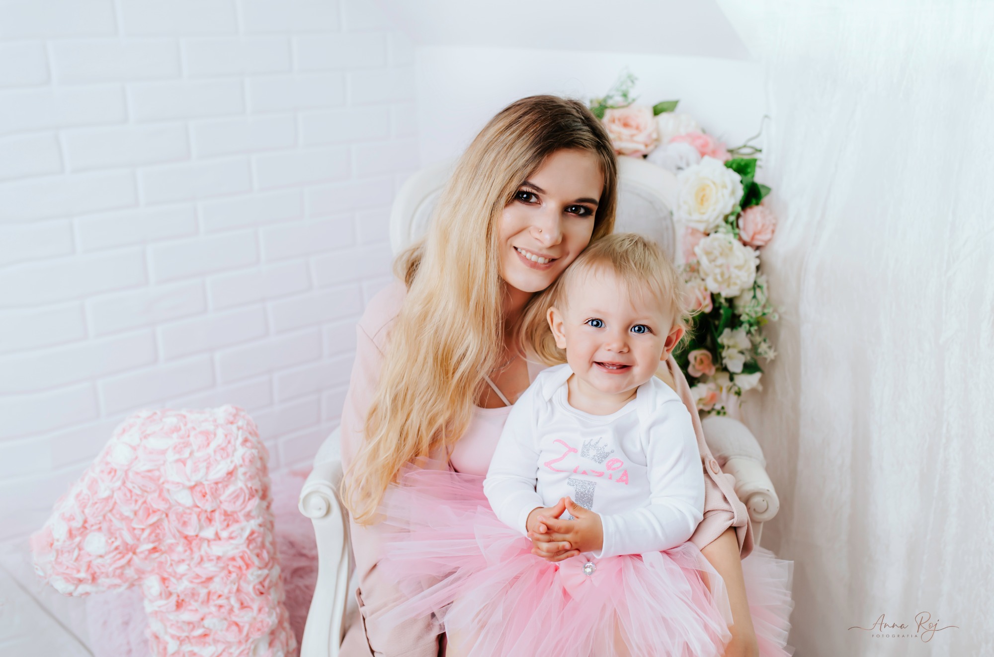 Uśmiechnięta mama z blond włosami i mała dziewczynka w różowej tiulowej spódniczce siedząca na białym krześle, sesja urodzinowa z dekoracją kwiatową w tle