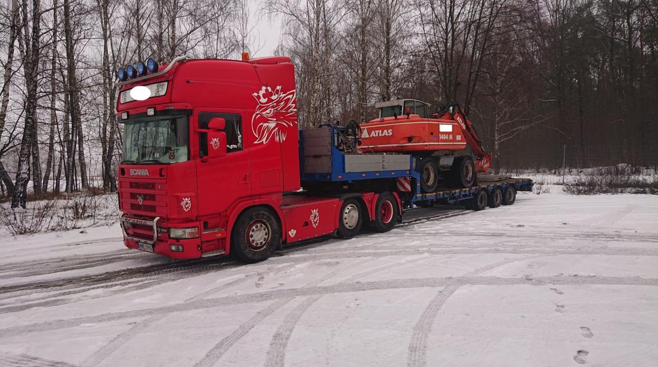 Czerwona ciężarówka Scania z białym orłem na kabinie przewozi pomarańczową koparkę Atlas na niskopodwoziowej naczepie w zimowej scenerii.