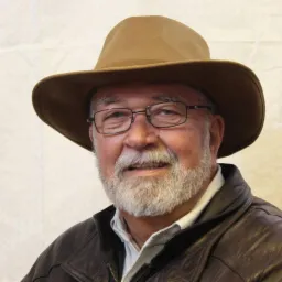 Portret uśmiechniętego starszego mężczyzny z siwą brodą, w okularach i brązowym kapeluszu, ubrany w skórzaną kurtkę.