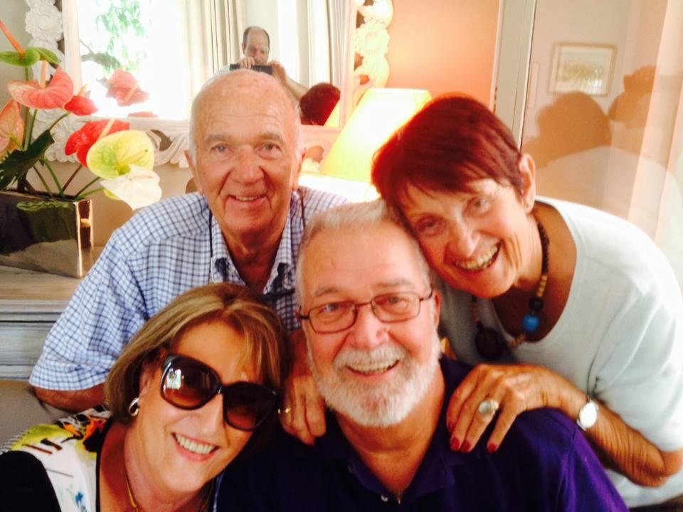 Grupa czterech uśmiechniętych, starszych osób pozuje do selfie, z mężczyzną robiącym zdjęcie widocznym w lustrze w tle, obok dekoracyjnych kwiatów anturium.