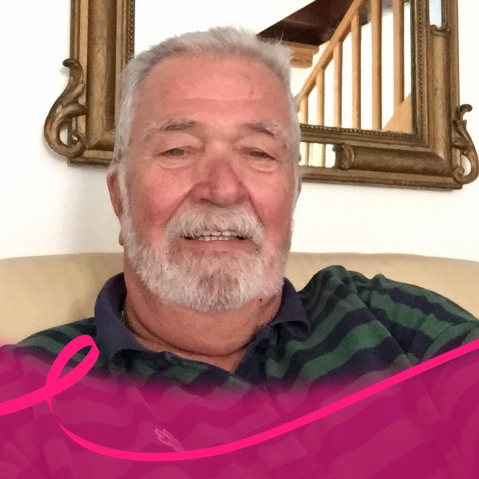 Portret uśmiechniętego mężczyzny z siwą brodą w zielono-granatowej koszulce w paski, z dekoracyjnym złotym lustrem w tle i różową wstążką na pierwszym planie.