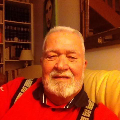 Portret starszego mężczyzny z białą brodą w czerwonej koszuli z szelkami, siedzącego na żółtej kanapie na tle regału z książkami i obrazu.