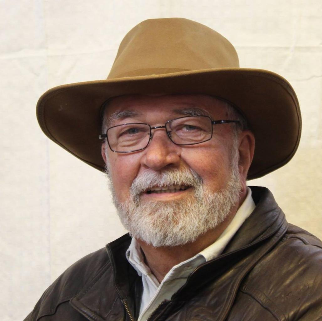 Portret uśmiechniętego starszego mężczyzny z siwą brodą, w okularach i brązowym kapeluszu, ubrany w skórzaną kurtkę.