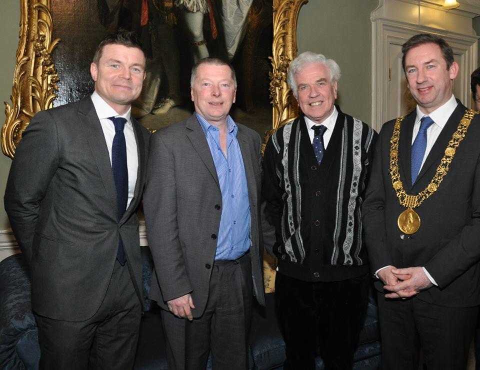 Grupa czterech mężczyzn w garniturach pozuje do zdjęcia w eleganckim wnętrzu z obrazem w złotej ramie w tle, jeden z nich ma na sobie łańcuch z medalionem.