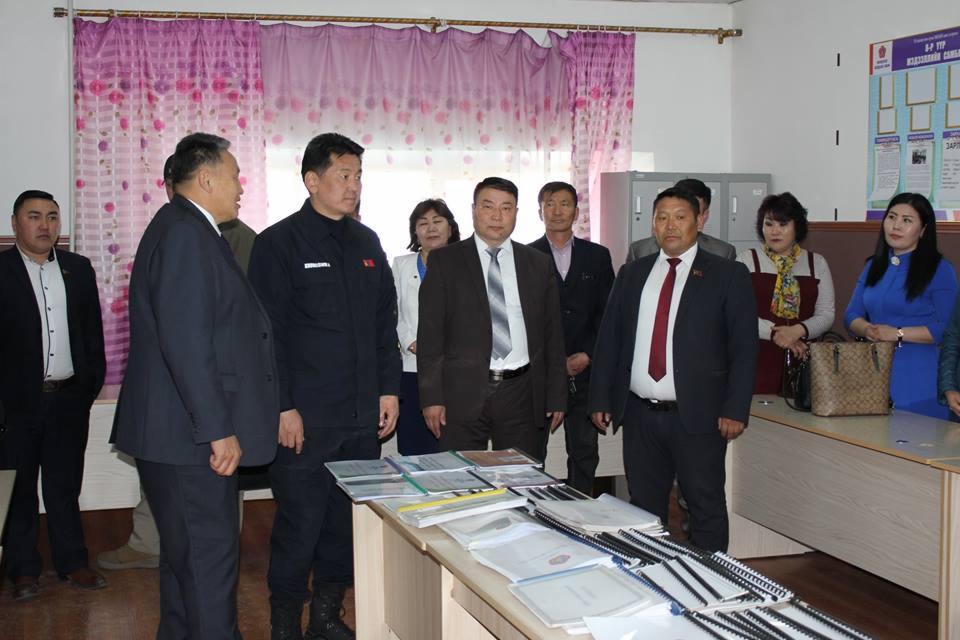 Grupa osób w formalnych strojach stoi za stołem z dokumentami i segregatorami w jasnym pomieszczeniu z zasłonami w kwiaty.