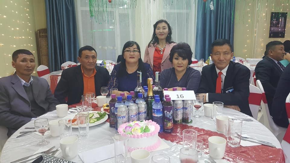 Grupa osób siedzi przy stole podczas uroczystości, na stole napoje, przekąski i tort z numerem 16.