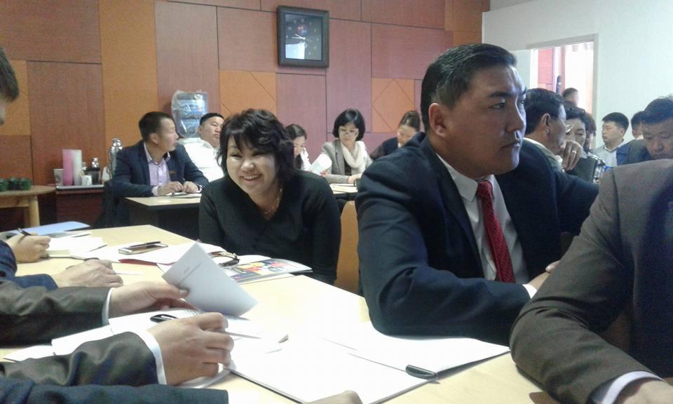 Grupa osób uczestniczy w spotkaniu biznesowym przy stołach, część z nich w garniturach, inni notują lub przeglądają dokumenty, w tle widoczny dystrybutor wody i zegar na ścianie.