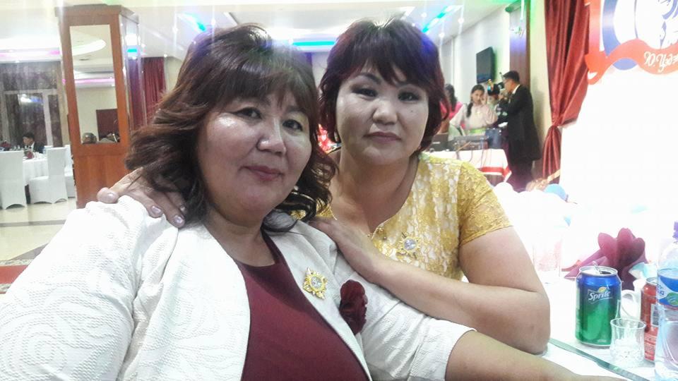 Dwie uśmiechnięte kobiety pozują do zdjęcia w sali bankietowej z dekoracjami i stołami w tle. Jedna ma na sobie kremową marynarkę z ozdobną broszką, a druga żółtą koronkową bluzkę.