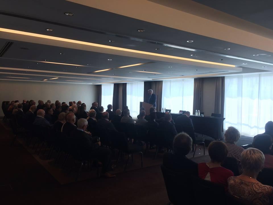 Sala konferencyjna z rzędami siedzących osób słuchających prelegenta za mównicą, jasne światło wpadające przez okna z zasłonami w tle.