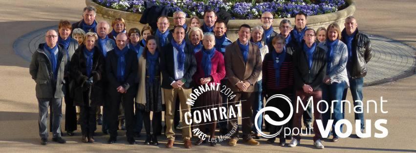 Grupa osób w różnym wieku, ubranych w niebieskie szale, pozuje do zdjęcia przed fontanną z fioletowymi kwiatami, z widocznym logo i napisem 'Mornant pour Vous'.