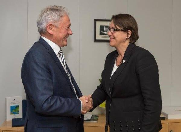 Uśmiechnięty mężczyzna w granatowym garniturze i kobieta w czarnym żakiecie uściskują sobie dłonie na tle szarej ściany z obrazkiem i drewnianego biurka.