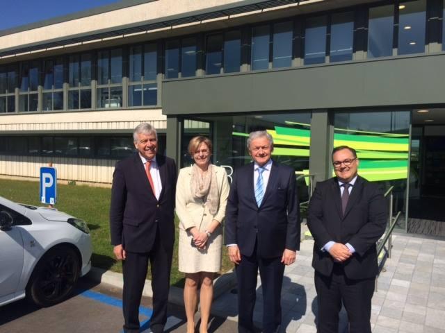 Grupa czterech osób w garniturach, pozujących przed nowoczesnym budynkiem z zielonymi akcentami, obok zaparkowanego białego samochodu z niebieskim znakiem parkingu.