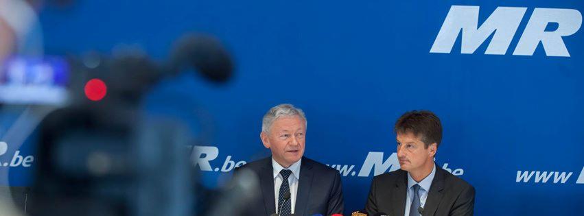 Dwóch mężczyzn w garniturach siedzi przy stole podczas konferencji prasowej z logo MR w tle.