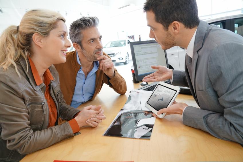 Klientka z partnerem rozmawiają z doradcą finansowym w salonie samochodowym, analizując dane na tablecie i wydrukowane materiały.