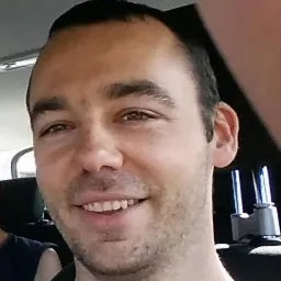 Uśmiechnięty mężczyzna w busie, widoczny fragment zagłówka i uchwyt. Portret wewnątrz pojazdu, prawdopodobnie podczas podróży.