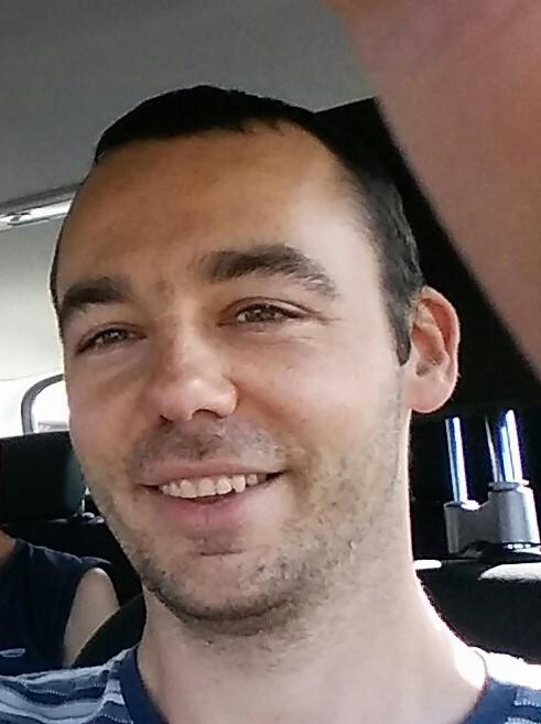 Uśmiechnięty mężczyzna w busie, widoczny fragment zagłówka i uchwyt. Portret wewnątrz pojazdu, prawdopodobnie podczas podróży.