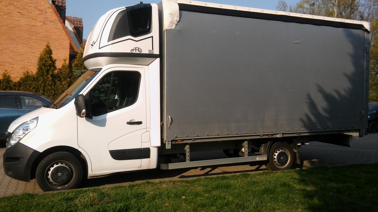 Biały bus dostawczy z szarą plandeką, zaparkowany na trawniku obok chodnika, z widocznym logo firmy na kabinie sypialnej.