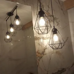 Zawieszone czarne lampy w geometrycznych kloszach z żar&oacute;wkami na tle ściany wyłożonej marmurowymi płytkami z brązowymi żyłami, odbicie lamp w lustrze.