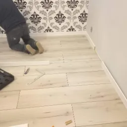 Pracownik na kolanach układa jasne panele podłogowe, widoczne narzędzia: nóż, kątownik i obcinarka do listew, ściana z tapetą w ornamenty.
