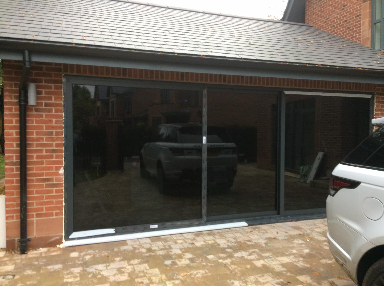 Nowoczesne, przesuwne drzwi garażowe z ciemnego szkła, z widocznym samochodem wewnątrz, osadzone w ceglanej elewacji budynku z szarym dachem.