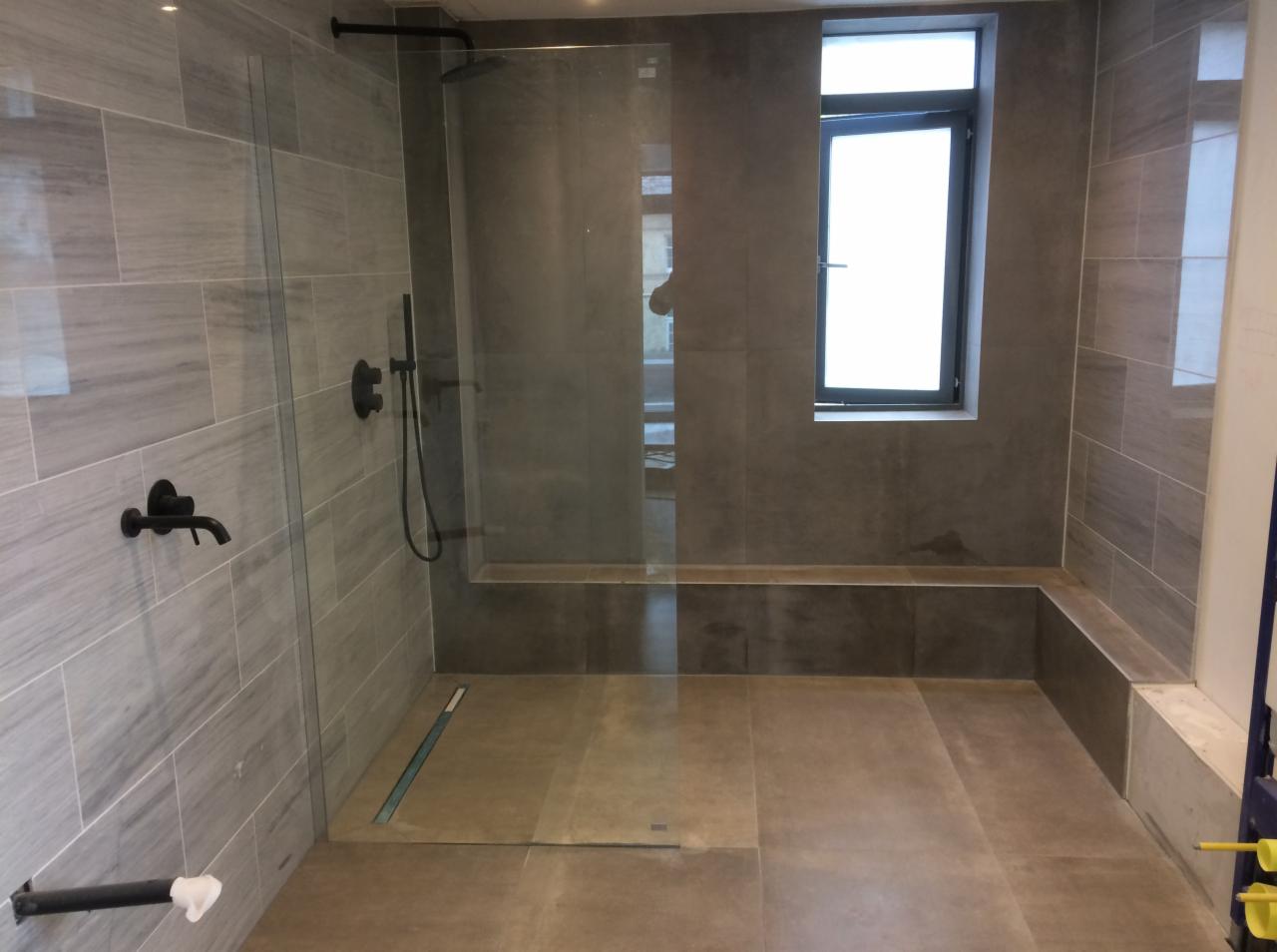 Nowoczesna łazienka z szarymi płytkami imitującymi marmur, kabiną prysznicową typu walk-in z czarną armaturą i odpływem liniowym w podłodze, małe okno.