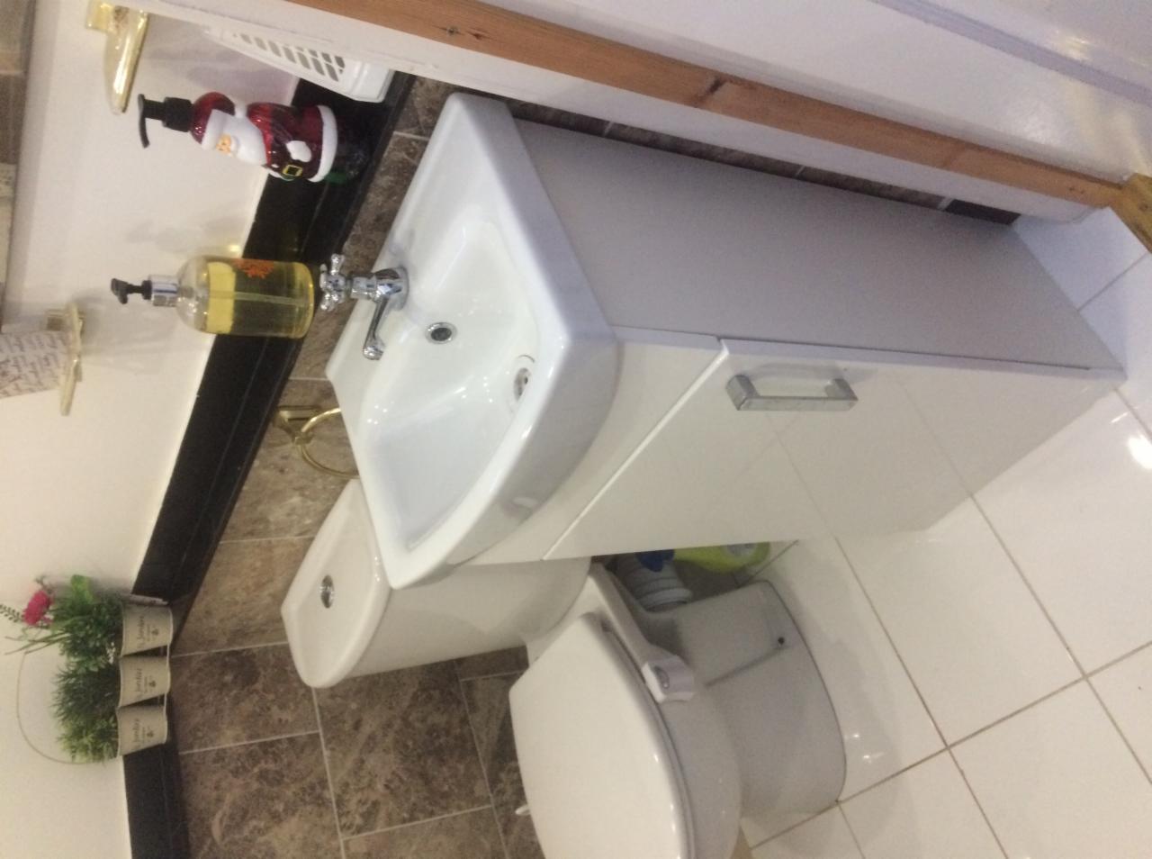 Biała umywalka z szafką, toaleta i bidet w łazience z brązowymi płytkami na ścianie i białymi na podłodze, z dekoracyjnym dozownikiem mydła i figurką Świętego Mikołaja na półce.