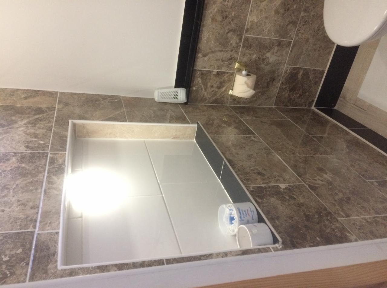 Błyszczące, białe płytki ułożone w brodziku kabiny prysznicowej, otoczone marmurową mozaiką na ścianach i podłodze, z widocznym dozownikiem mydła i rolką papieru toaletowego.