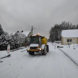 Żółta wywrotka na ośnieżonej drodze w terenie zabudowanym, widoczne ślady opon, biało-czerwone słupki ograniczające pas ruchu, ponure niebo.
