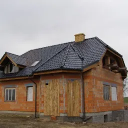Dom w trakcie budowy z ciemną dachówką, widoczna murowana konstrukcja, drewniane zabezpieczenia okien i otwarta przestrzeń poddasza, ceglany komin.