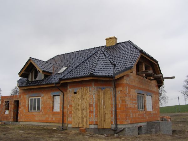 Dom w trakcie budowy z ciemną dachówką, widoczna murowana konstrukcja, drewniane zabezpieczenia okien i otwarta przestrzeń poddasza, ceglany komin.
